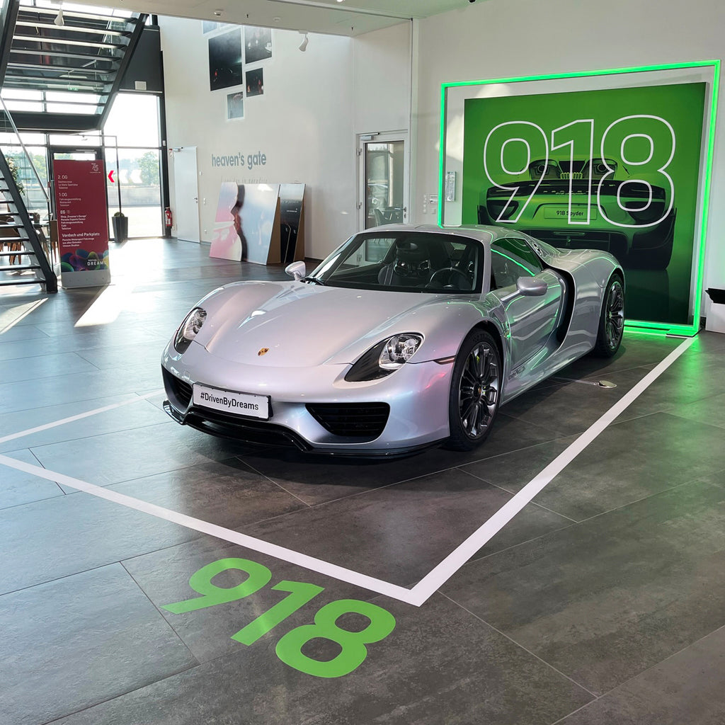 Ausstellung eines Porsche 918 Spyder in einem Showroom, hervorgehoben durch eine grüne LED-Leuchtkontur von Modulap im Hintergrund und das Branding #DrivenByDreams. Der Sportwagen steht in einem markierten Bereich mit der Nummer 918, passend zur Modellbezeichnung.