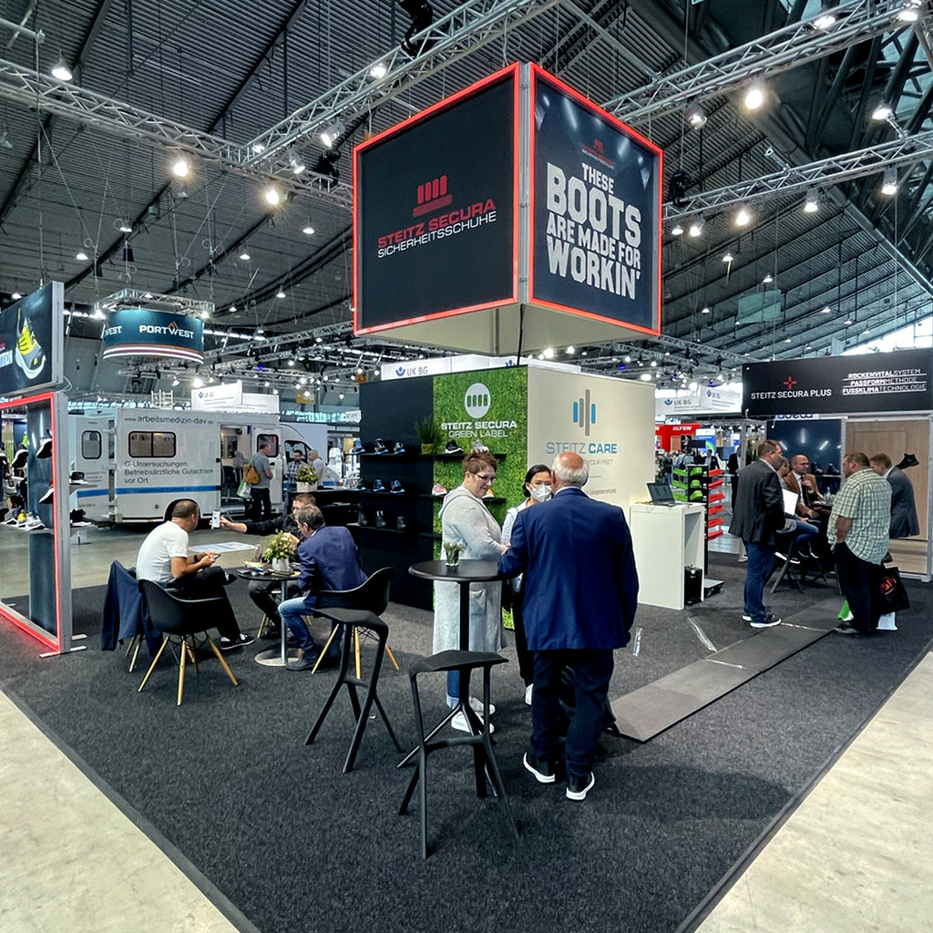Messestand von Steitz Secura auf einer Fachmesse, präsentiert Sicherheits- und Arbeitsschuhe. Der Stand hebt sich durch moderne Displays, eine große beleuchtete Würfelstruktur mit Branding und Bereiche für Gespräche und Produktpräsentationen hervor.