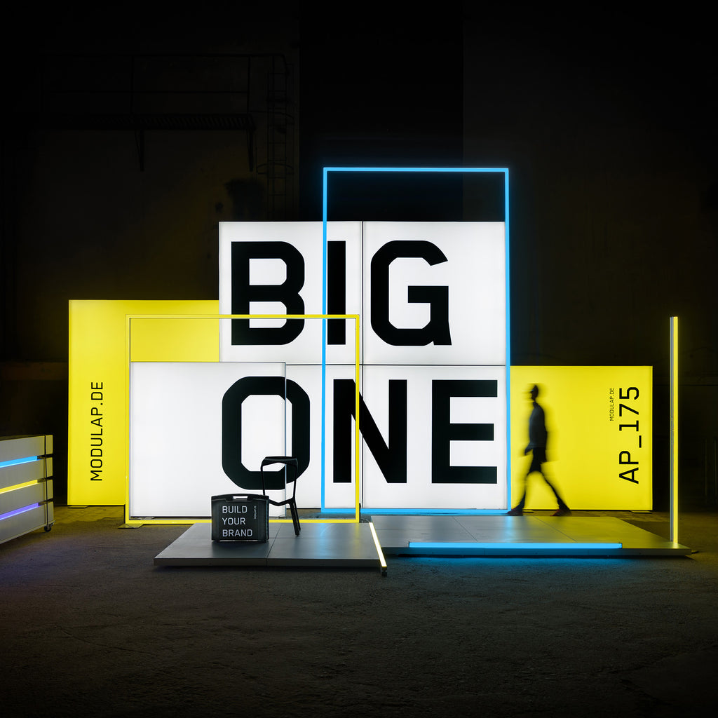 Modularer Messestand von Modulap mit auffälligen LED-Leuchtkonturen in Gelb und Blau. Große, beleuchtete Lightboxes mit der Aufschrift 'BIG ONE' dominieren das Design und vermitteln eine starke Markenbotschaft. Der Stand kombiniert klare Linien und moderne Präsentationselemente.