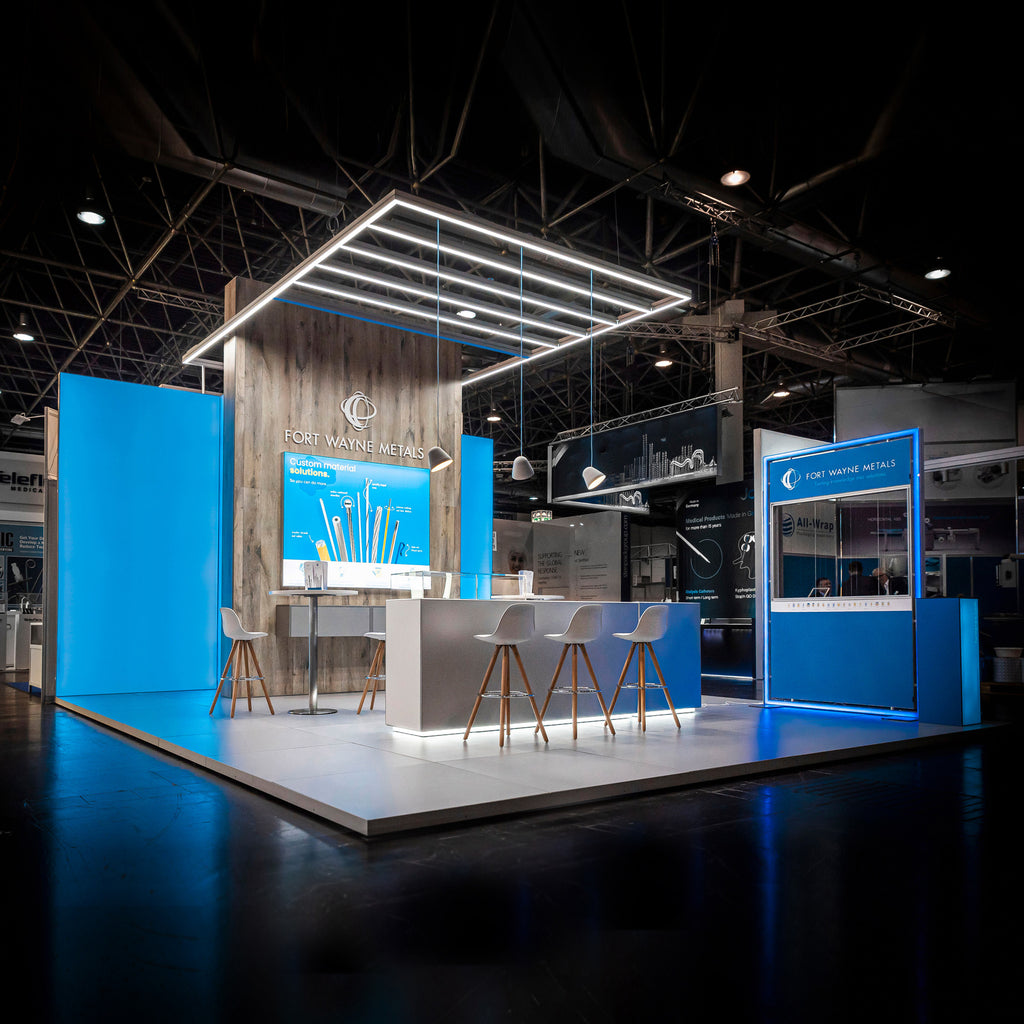 Messestand von Fort Wayne Metals mit modernem Design, LED-Beleuchtung und blauen Akzentelementen. Der Stand kombiniert Holzwände, elegante Sitzgelegenheiten und eine beleuchtete Deckenstruktur, die die Marke professionell und einladend präsentiert.