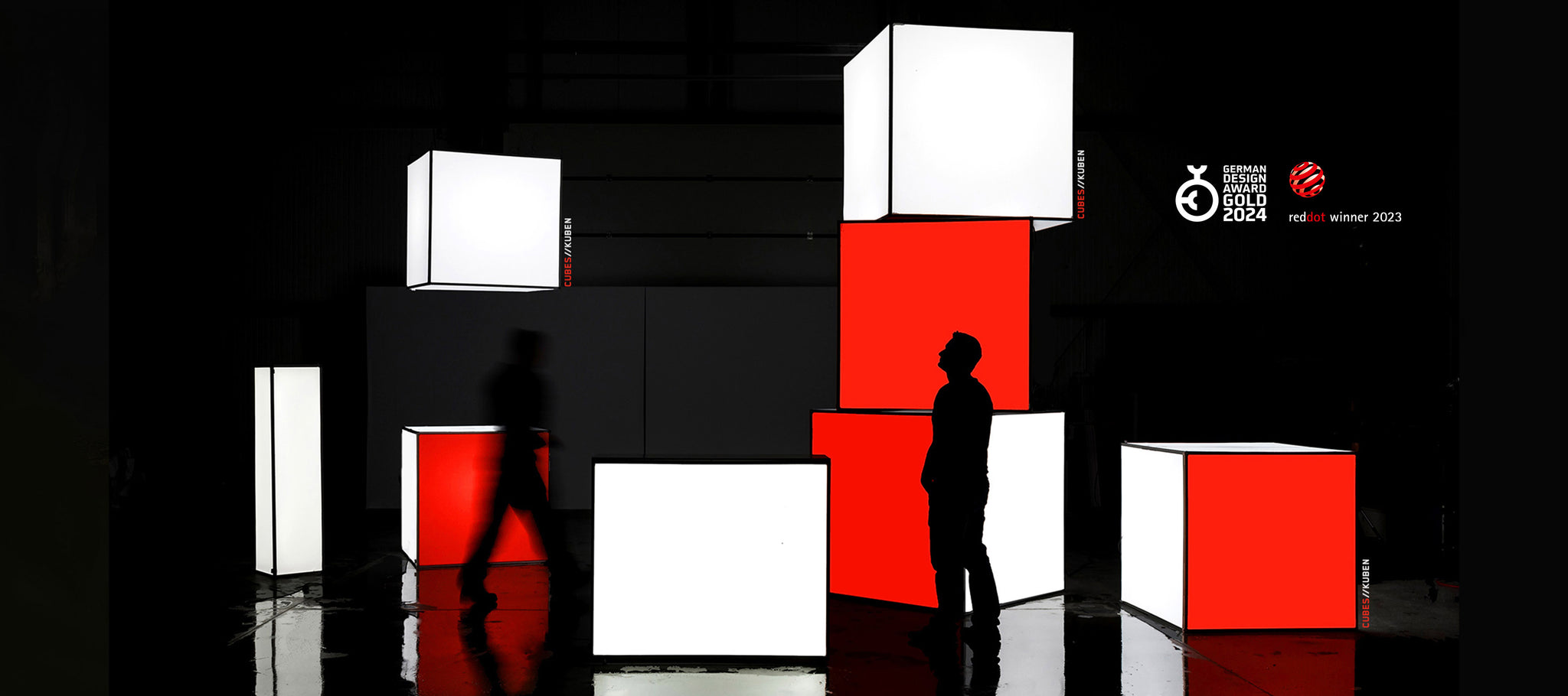 Messestand mit mehreren LED-Leuchtwürfeln in Weiß und Rot, die in verschiedenen Größen und Höhen angeordnet sind. Zwei Personen bewegen sich durch den Stand, der eine dynamische und auffällige Präsentationsfläche bietet. Die Leuchtwürfel sind strategisch platziert und schaffen eine moderne und visuell ansprechende Umgebung, ideal für Markenpräsentationen und interaktive Ausstellungen.