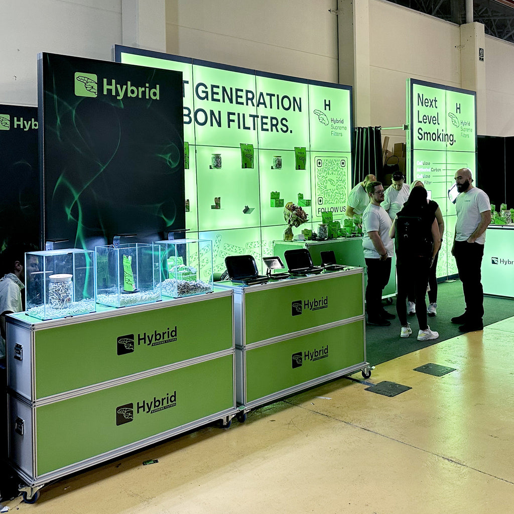 Messestand von Hybrid mit Modulap Cases als Präsentationsfläche. Die Cases dienen als stilvolle Podeste für Glasvitrinen, die Produkte ausstellen. Im Hintergrund ist eine grüne, beleuchtete LED-Wand mit dem Slogan 'Next Generation Carbon Filters' zu sehen. Moderne Gestaltung für effektive Produktpräsentation.