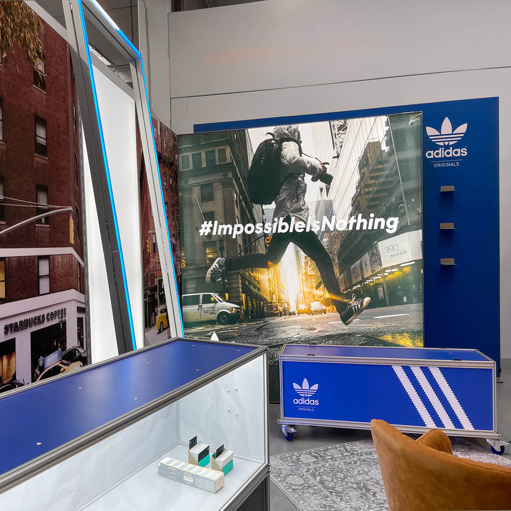 Adidas Originals Messestand mit beleuchteter Werbefläche und dem Slogan '#ImpossibleIsNothing'. Der Stand kombiniert moderne LED-Rahmen, eine beleuchtete Vitrine und einen Flightcase Counter im markentypischen Design, um die dynamische Markenpräsenz zu betonen.