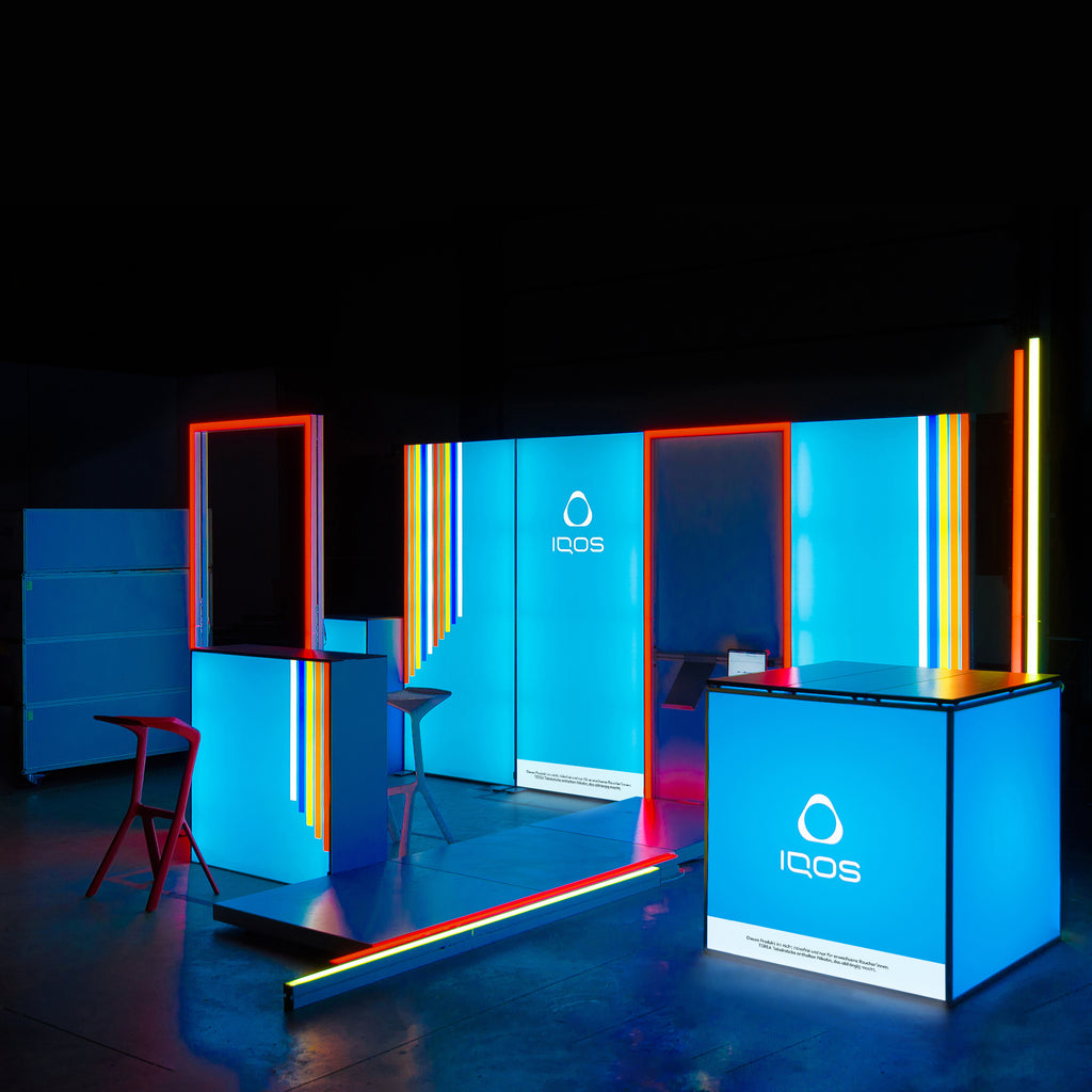 Modularer Messestand von IQOS mit leuchtenden LED-Elementen und Lightboxes in Blau, kombiniert mit roten und gelben Akzenten. Das moderne Design schafft eine dynamische und futuristische Präsentationsfläche für die Marke.