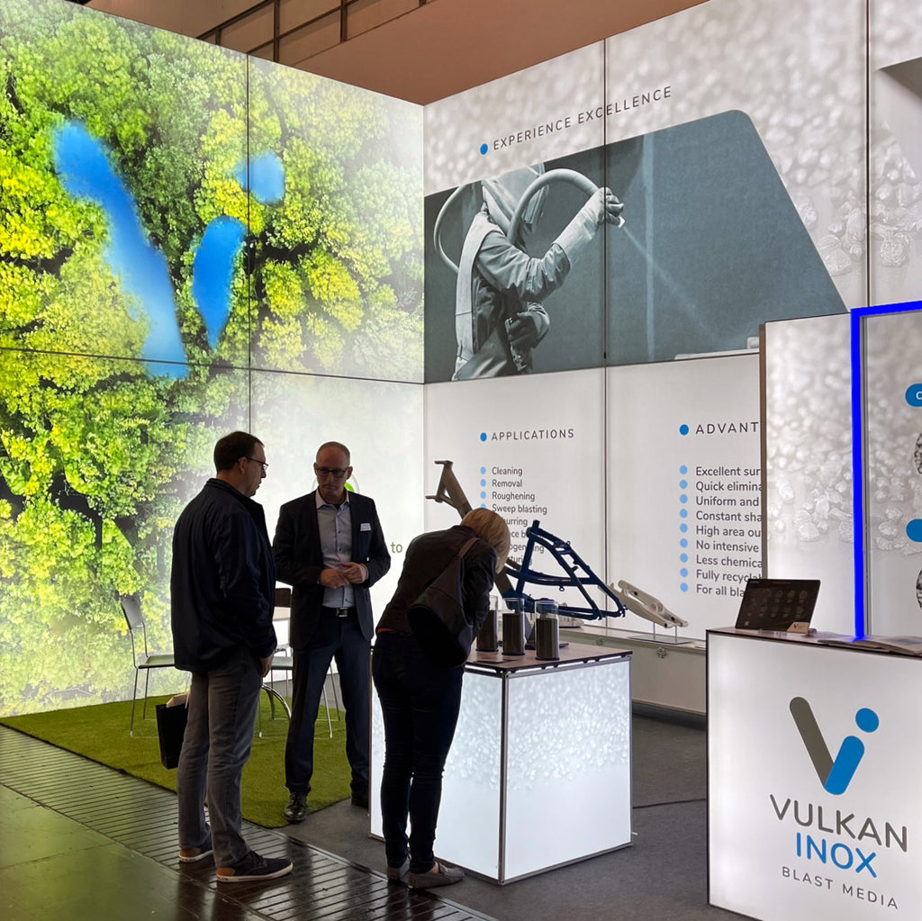 Messestand von Vulkan Inox mit großen, beleuchteten Lightboxes, die Anwendungen und Vorteile der Blast Media Produkte präsentieren. Der Stand kombiniert beeindruckende visuelle Hintergründe mit informativen Displays und interaktiven Produktpräsentationen.