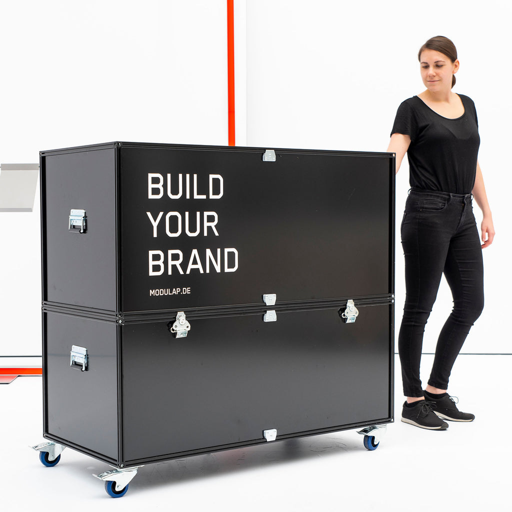 Modulap Transport Case mit 'Build Your Brand' Aufdruck, gezeigt mit einer Person zur Größendarstellung. Das robuste Case auf Rollen bietet sicheren Transport und flexible Einsatzmöglichkeiten auf Messen und Events.