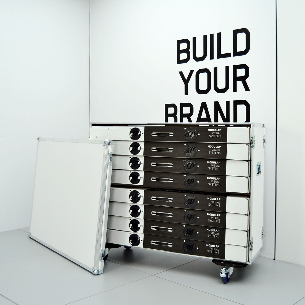 Modulap Stackable Cases: Hochwertige, stapelbare Transportcases von Modulap Visual Systems, die als modulare Lösung für den einfachen Transport und die Aufbewahrung von Messe- und Displayelementen dienen. Mit dem Slogan 'Build Your Brand' im Hintergrund.